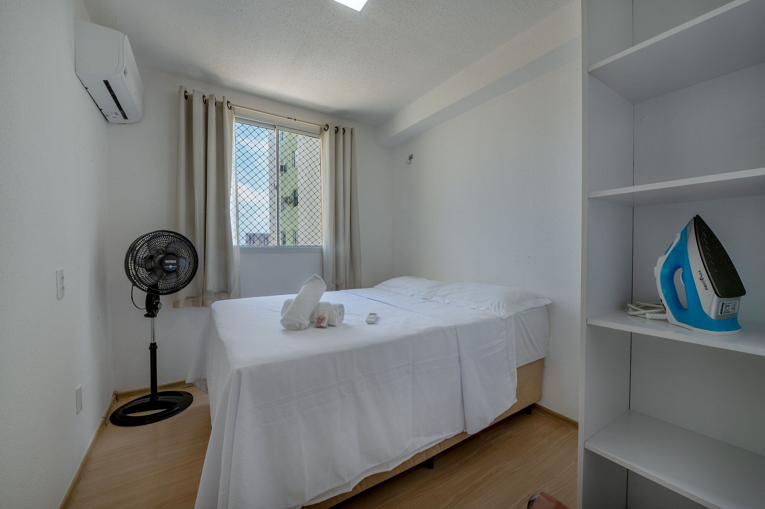 Una habitación pequeña y limpia con cama, ventilador, ventana con cortinas y una estantería con plancha.