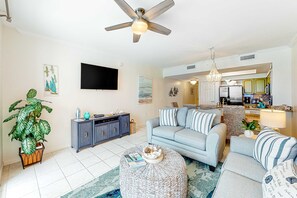 A modern living room with a grey sofa, a coffee table, and a TV. There is a kitchen visible in the background. - Majestic Sun B1010 | Beachfront Bliss with Pool! (Miramar Beach)