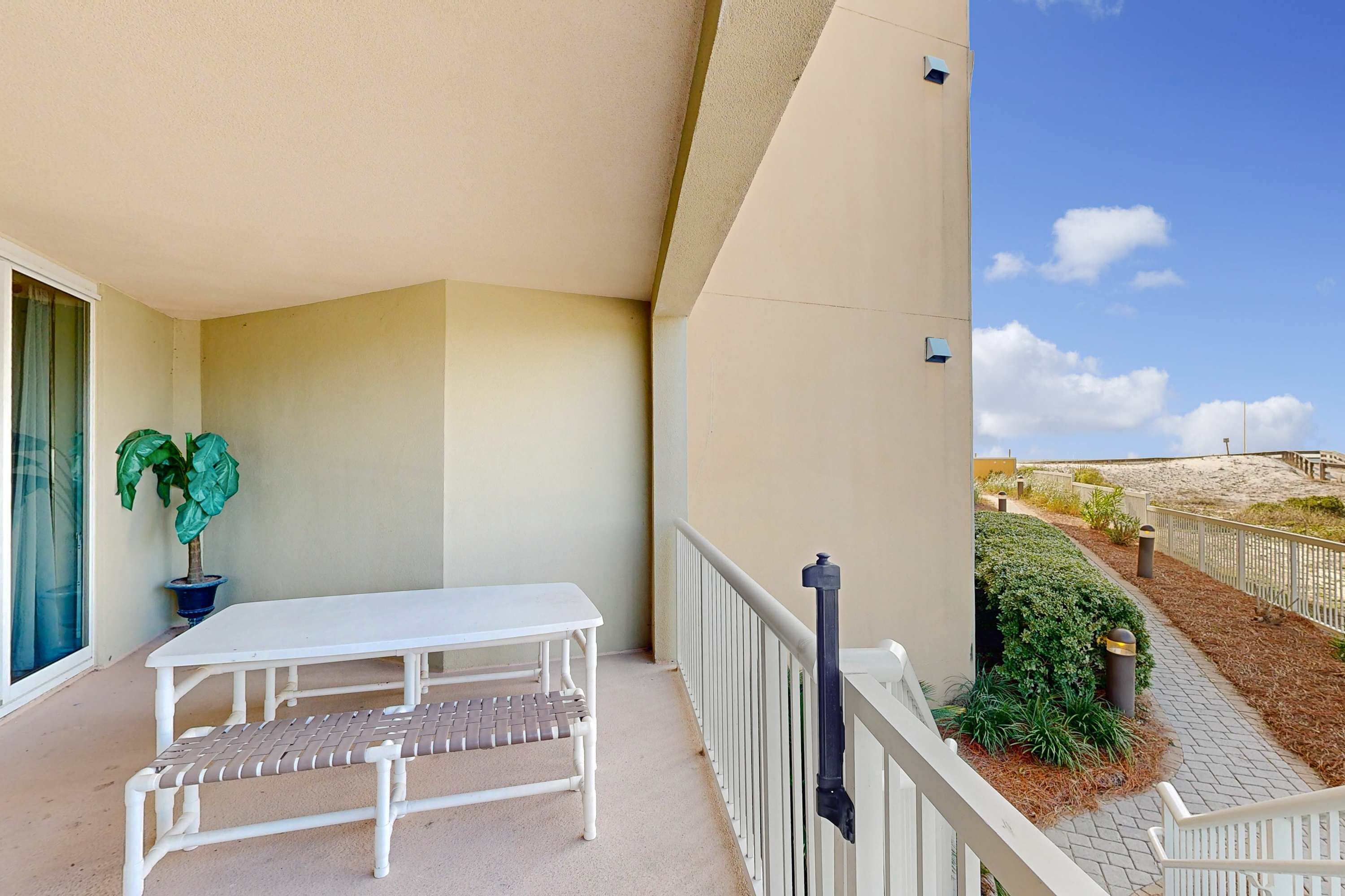 A balcony with a white table and bench, a potted plant, and a view of a sandy beach area.