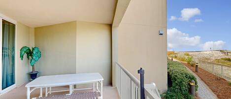 A balcony with a white table and bench, a potted plant, and a view of a sandy beach area.