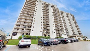 A high-rise hotel with multiple balconies, a parking lot, and a clear sky. - Second Floor Condo with Shared Pool, Partial Ocean View, Washer/Dryer, and WiFi (Miramar Beach)