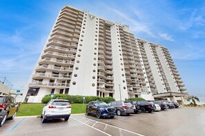 A high-rise hotel with multiple balconies, a parking lot, and a clear sky. - Second Floor Condo with Shared Pool, Partial Ocean View, Washer/Dryer, and WiFi (Miramar Beach)