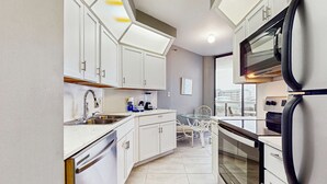 A modern kitchen with white cabinets, a dishwasher, a sink, a refrigerator, and an oven. There is a dining table with chairs and a window providing a view of the city. - Second Floor Condo with Shared Pool, Partial Ocean View, Washer/Dryer, and WiFi (Miramar Beach)