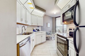 A modern kitchen with white cabinets, a dishwasher, a sink, a refrigerator, and an oven. There is a dining table with chairs and a window providing a view of the city. - Second Floor Condo with Shared Pool, Partial Ocean View, Washer/Dryer, and WiFi (Miramar Beach)