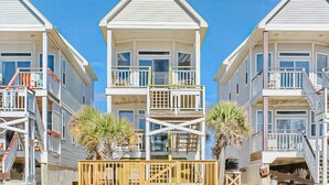 Une rangée de maisons situées en bord de mer, dotées de balcons et de terrasses en bois, sous un ciel d’un bleu limpide.