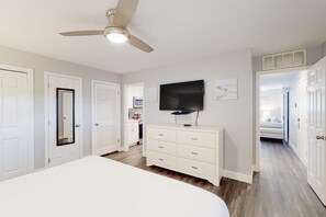 A bedroom with a bed, a ceiling fan, a TV, a dresser, and a door leading to a bathroom. - Ground-floor condo near beach with WiFi, central ac, patio, & onsite pool,tennis (Miramar Beach)
