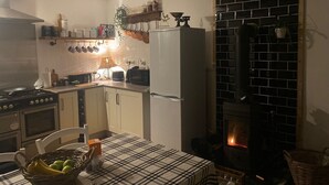 Una cocina con estufa, refrigerador y estufa de leña. Hay una mesa con mantel a cuadros y una cesta de fruta.
