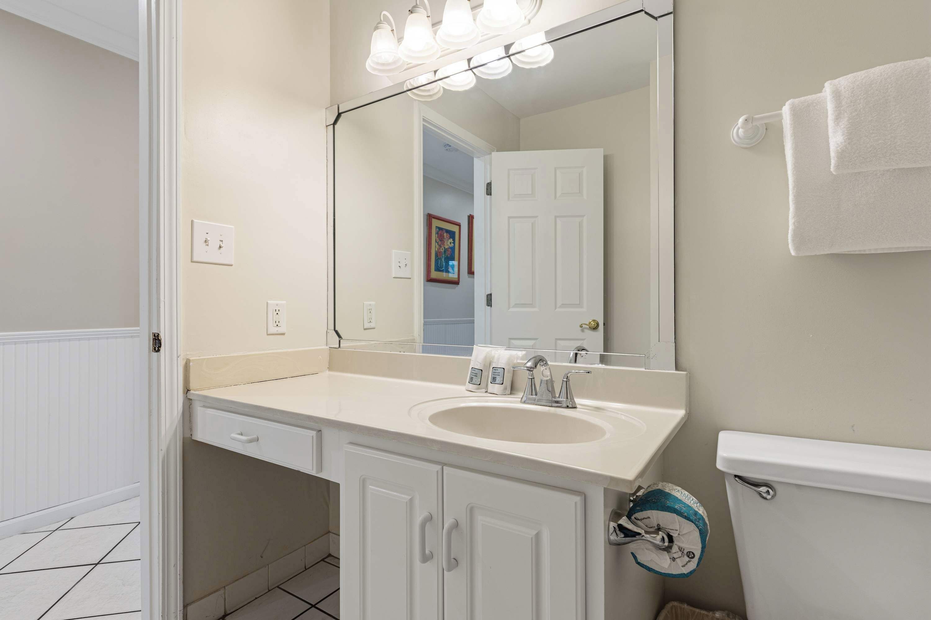 A bathroom with a white sink, a mirror, and a toilet. There are lights above the mirror and a towel rack on the wall.