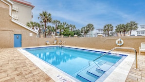Piscina retangular com borda azulejada azul, rodeada por um pátio de tijolos. Há cadeiras de descanso e um salva-vidas preso à borda da piscina.