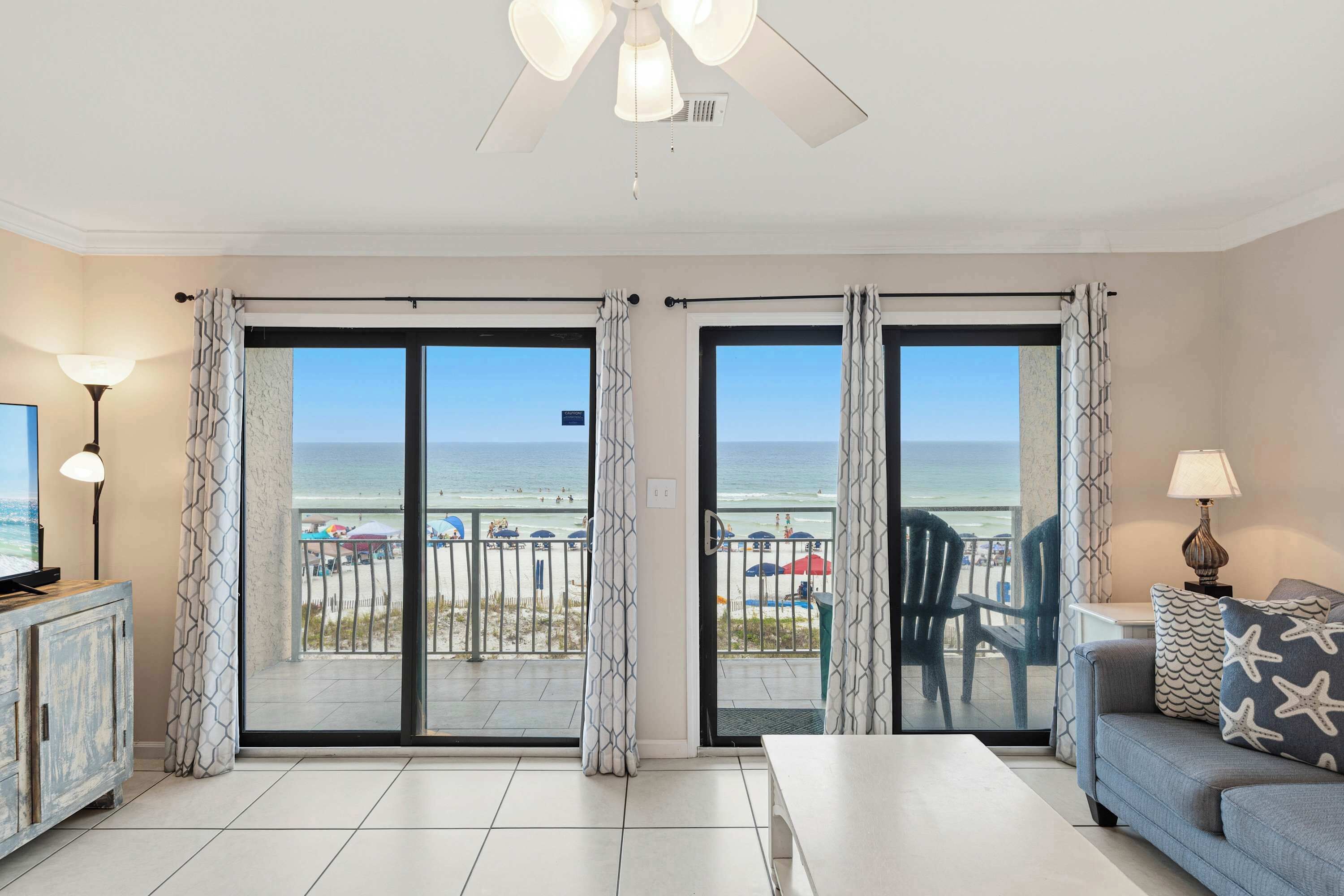 Un salon avec vue sur la plage, un ventilateur de plafond et un canapé agrémenté de coussins en forme d’étoiles de mer.