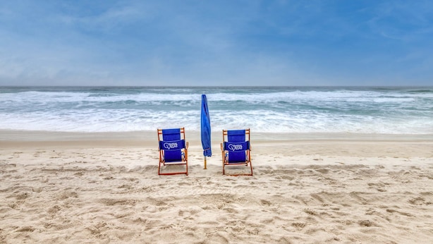 Duas cadeiras de praia com toalhas azuis numa praia arenosa, viradas para o oceano.