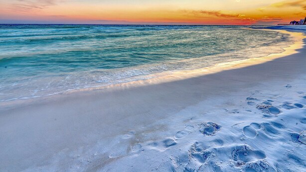Ein Strand mit Fußspuren im Sand und einem Sonnenuntergang im Hintergrund.