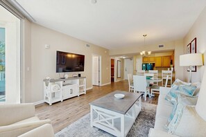 A spacious living room with a TV, white furniture, and a dining area in the background. - Gorgeous coastal condo with shared pool and private balcony (Destin)