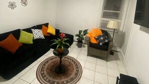Ein Wohnzimmer mit schwarzem Sofa, Couchtisch, Esstisch und Fernseher an der Wand.