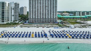 Um hotel na praia com um edifício grande, uma praia arenosa com guarda-sóis e uma vista clara do oceano.