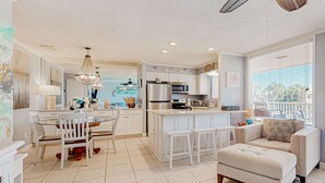 A modern kitchen with white cabinetry, a central island, and a dining area with a glass table and white chairs. - 2nd-floor condo with beach access, pool, tennis courts, & on-site laundry (Destin)