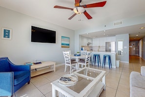 A modern living room with a ceiling fan, a flat-screen TV, a dining area with white chairs, and a kitchen in the background. - Bright, Gulf view condo w balcony & shared lagoon pool&gym - steps to beach (Destin)