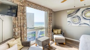 A modern room with a large window, a ceiling fan, a flat-screen TV, and a painting on the wall. - Ocean-view condo with pool, sauna, tennis courts & private beach access (Destin)