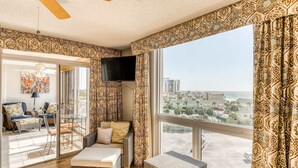 A spacious living room with a large window, wicker furniture, a flat-screen TV, and a city view. - Ocean-view condo with pool, sauna, tennis courts & private beach access (Destin)