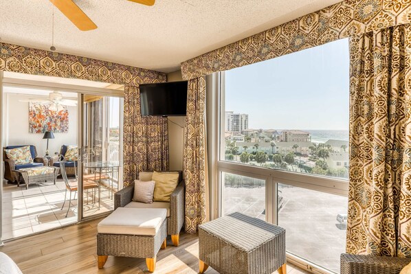 A spacious living room with a large window, wicker furniture, a flat-screen TV, and a city view. - Ocean-view condo with pool, sauna, tennis courts & private beach access (Destin)