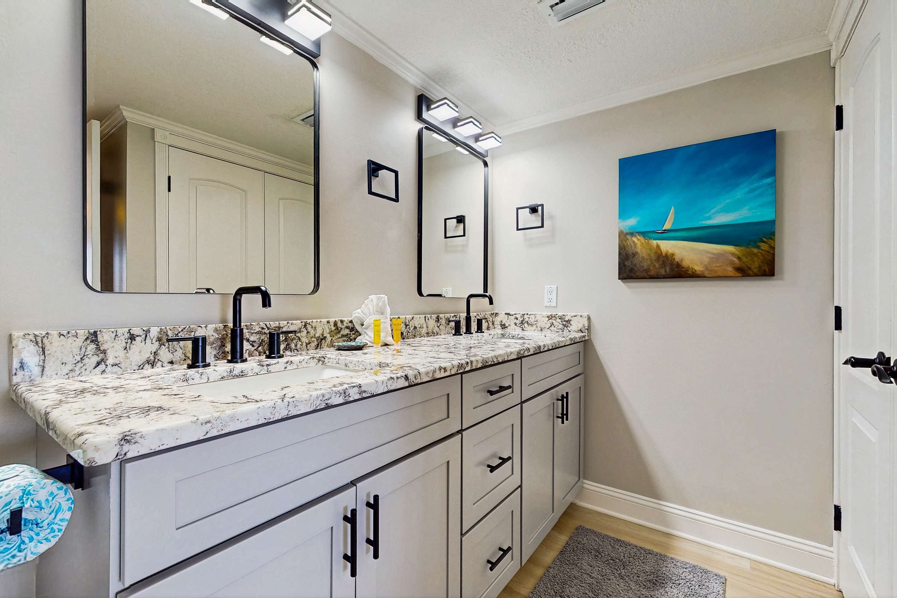 Une salle de bain avec un comptoir en marbre, des robinets noirs et un grand miroir. Un tableau représentant une scène de plage est accroché au mur.