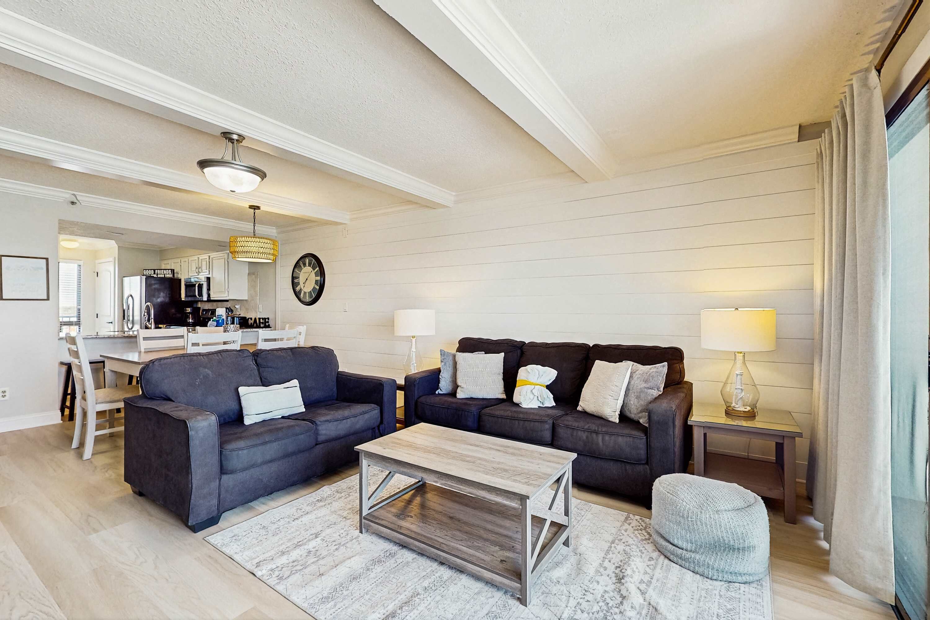 Ein modernes Wohnzimmer mit zwei dunklen Sofas, einem Holztisch und einem kleinen Teppich. Es gibt eine Essecke mit einem weißen Tisch und Stühlen, sowie eine Küche im Hintergrund.