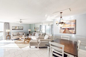 A spacious living room with a large sofa set, a dining table with chairs, a chandelier, and a large mirror reflecting the room. - Enchanting condo with tennis, pool, hot tub, balcony, & washer/dryer (Miramar Beach)