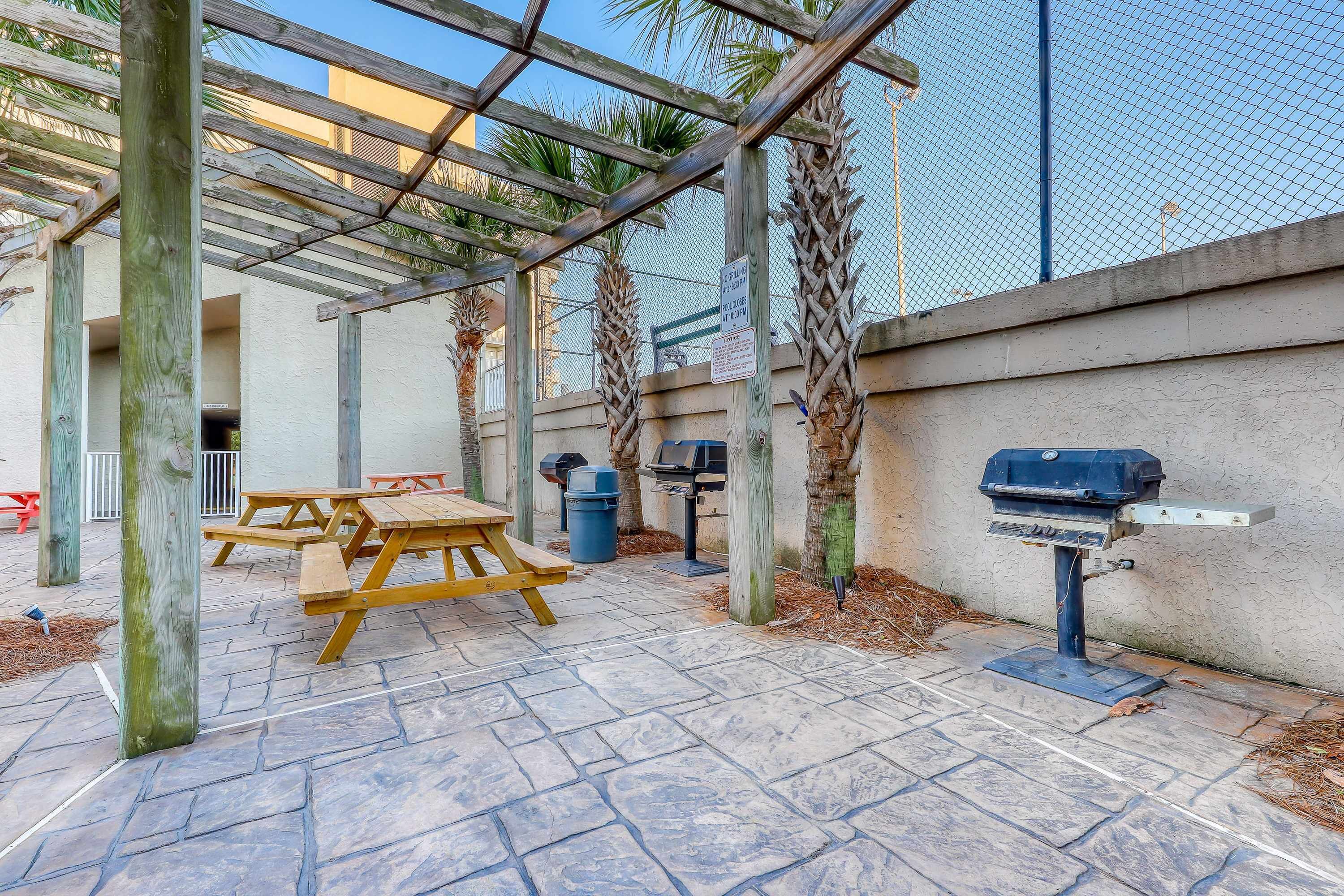 Ein Terrassenbereich im Freien mit einer Holzpergola, einem Picknicktisch, einem Grill und Mülleimern.