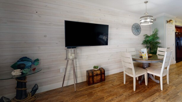 Een eetgedeelte met een ronde tafel, stoelen, een aan de muur bevestigde televisie, een klok en een potplant.