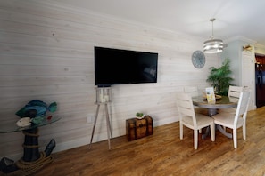 Un coin repas avec une table ronde, des chaises, un téléviseur fixé au mur, une horloge et une plante en pot.