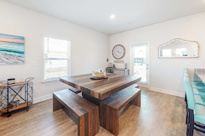 A spacious dining area with a wooden table and benches, a kitchen area with a clock and decorative items, and a view through the window. - Elegant Coastal Home w/Shared Pool, Tree-Lined View, 6-Minute Walk to the Beach (Port St. Joe)