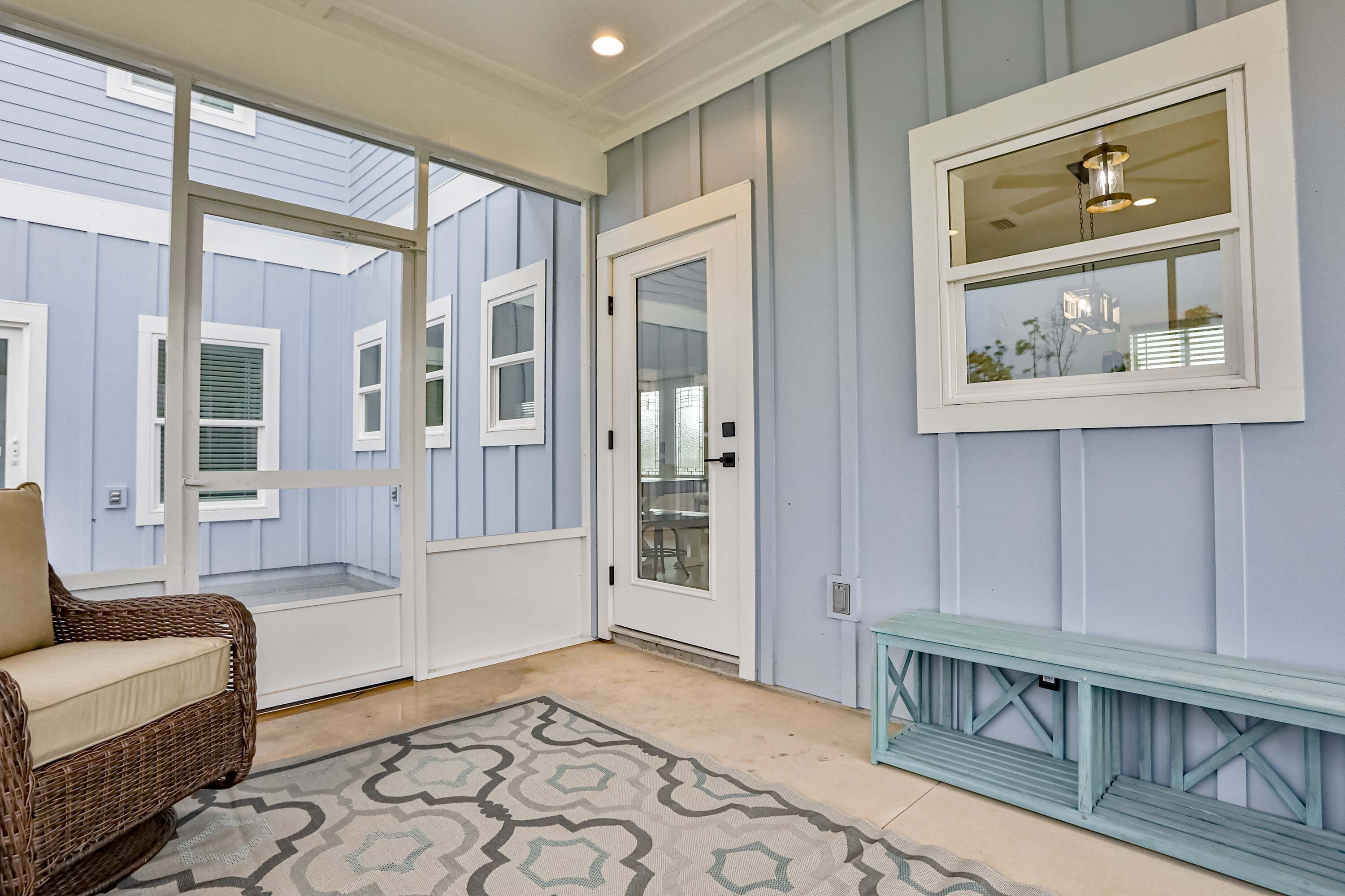 A room with a wicker chair, a patterned rug, a blue bench, and a wall-mounted mirror.