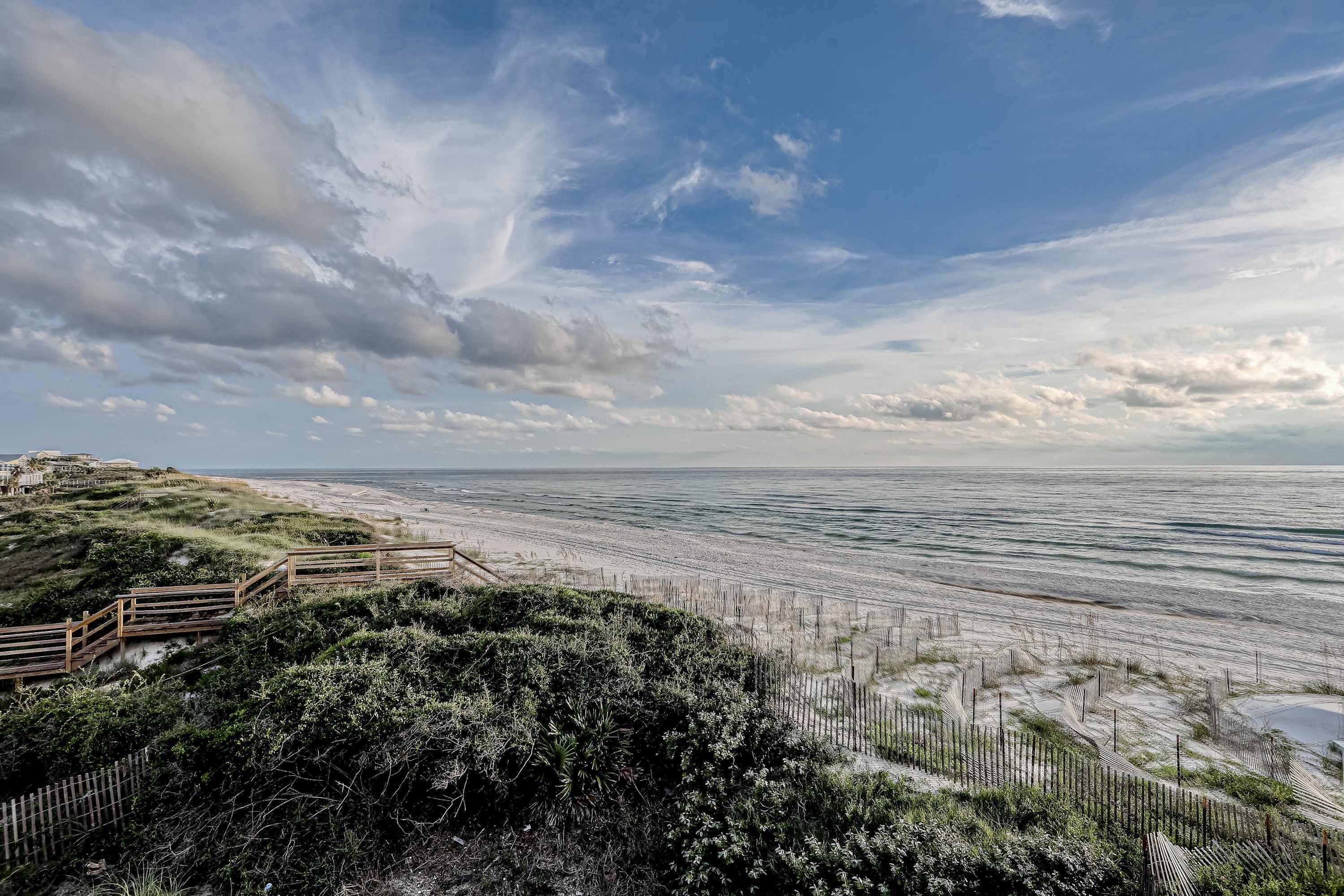 A coastal landscape with a wooden boardwalk leading to a sandy beach, surrounded by dunes and vegetation.