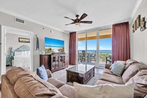 A spacious living room with a large sofa, a coffee table, and a television. There is a balcony with a view of the ocean. - Azure 202-Newly Refreshed 2026 (Fort Walton Beach)
