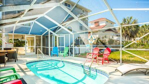 A covered outdoor swimming pool with a blue and white tiled edge, surrounded by red and green chairs, and a covered patio area with a view of a house and palm trees. - Beach Across the Street! Dog-Friendly Home with Private Pool, AC, WiFi & Games! (St. George Island)