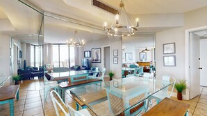 Un comedor espacioso con mesas y sillas de vidrio, un candelabro y vista al salón a través de un espejo grande.
