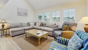 A living room with a sectional sofa, a coffee table, and a patterned armchair. There are two windows with blinds, a lamp on a side table, and a painting on the wall. - Waterfront villa with multi level home, shared pool & easy beach access (Destin)