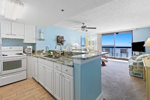 A modern kitchen with white cabinets, a granite countertop, and a built-in island. There is a ceiling fan, a large window with a view of the ocean, and a balcony with a TV. - 8th Floor Open Condo, On-site restaurant w bar, Multiple pools (Destin)