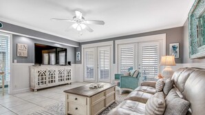 A living room with a white ceiling fan, a large TV, a white cabinet, a beige sofa, and a patterned rug.