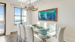 A dining area with a glass table, white chairs, a chandelier, and a framed painting. - Emerald Grande 1025 (Destin)