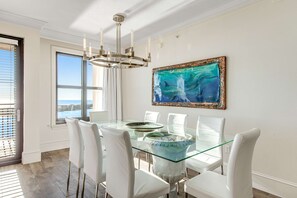 A dining area with a glass table, white chairs, a chandelier, and a framed painting. - Emerald Grande 1025 (Destin)