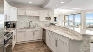 A modern kitchen with white cabinetry, a marble countertop, and a view of the ocean through large windows. - Emerald Grande 1025 (Destin)