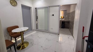 Ein modernes Zimmer mit Marmorboden, einem runden Tisch mit goldfarbener Basis, einem braunen Stuhl, einer Schiebetür aus Glas und einer kleinen Küchenzeile.