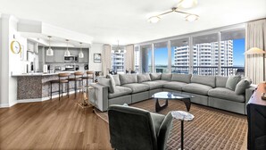 A modern living room with a large sectional sofa, a kitchen area, and large windows offering a city view. - Experience the perfect blend of luxury, comfort, and breathtaking Gulf views at Silver Shells (Destin)