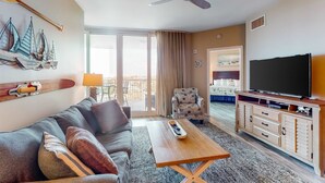 Habitación de hotel con sofá, mesa de centro, televisor sobre un mueble de madera, ventilador de techo y vistas al balcón exterior.