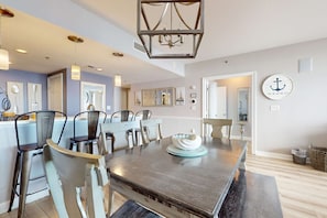 A dining area with a dark wooden table, light-colored chairs, and a chandelier. - Gorgeous beachfront condo w shared pool & beach access (Destin)