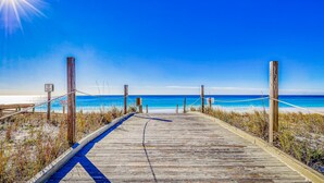 Une promenade en bois qui mène à une plage, avec un ciel bleu dégagé et l’océan à l’horizon.
