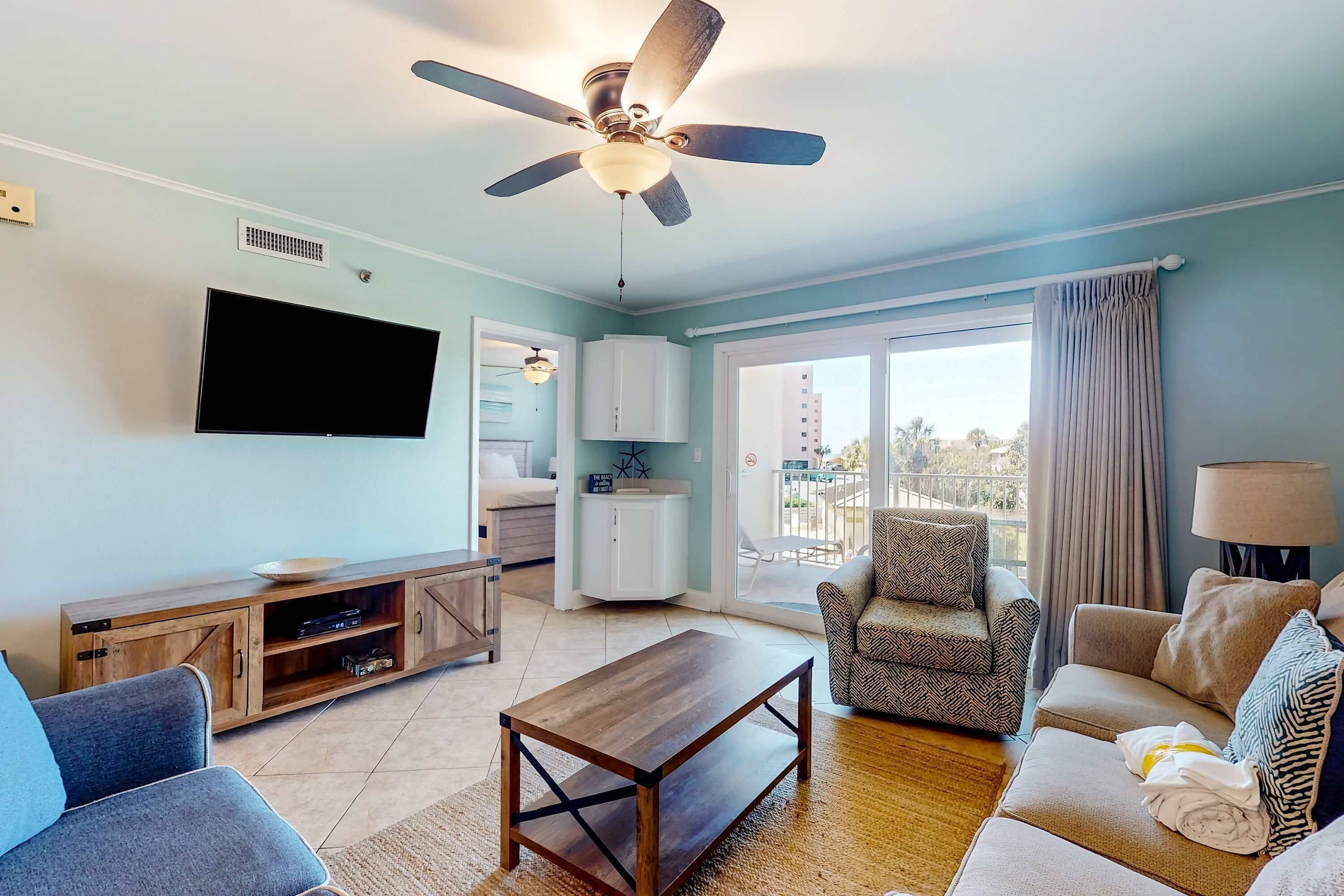 A living room with a ceiling fan, a flat-screen TV, a wooden cabinet, a sofa, a coffee table, and a balcony with a view.