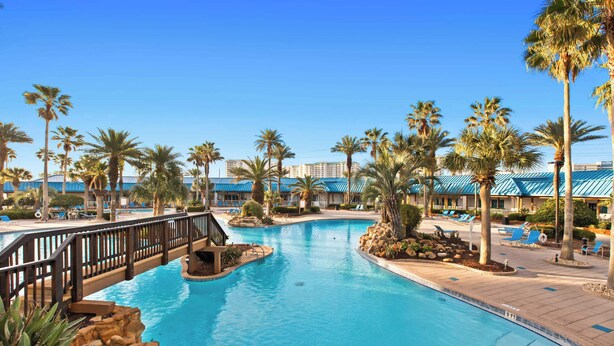 Zona de piscina en un resort, con palmeras, sillas de descanso y un puente.