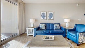 Una sala moderna con un sofá azul, dos mesitas de noche con lámparas, una mesa de centro con mandos a distancia y una obra de arte en la pared.